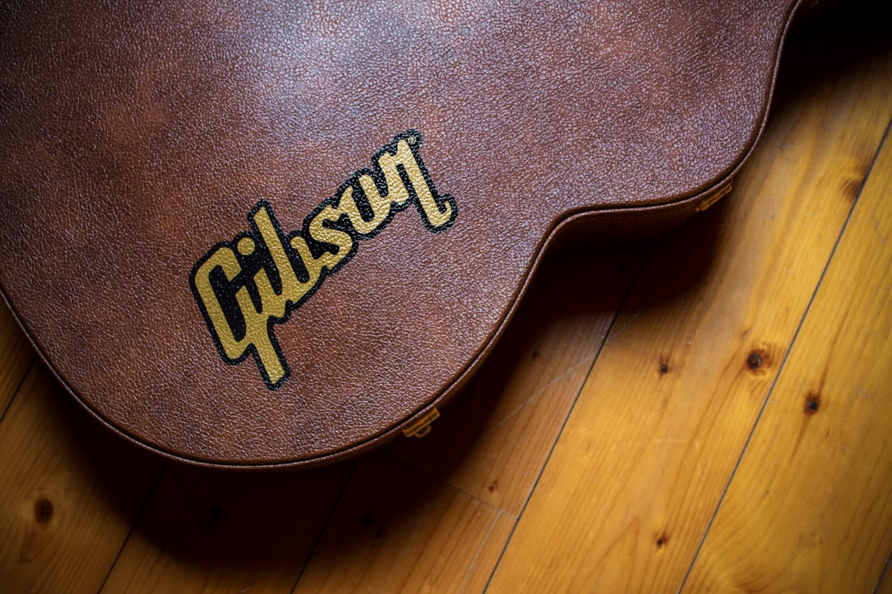 Home A close shot of a brown guitar case with 'Gibson' logo, placed on a wooden floor.